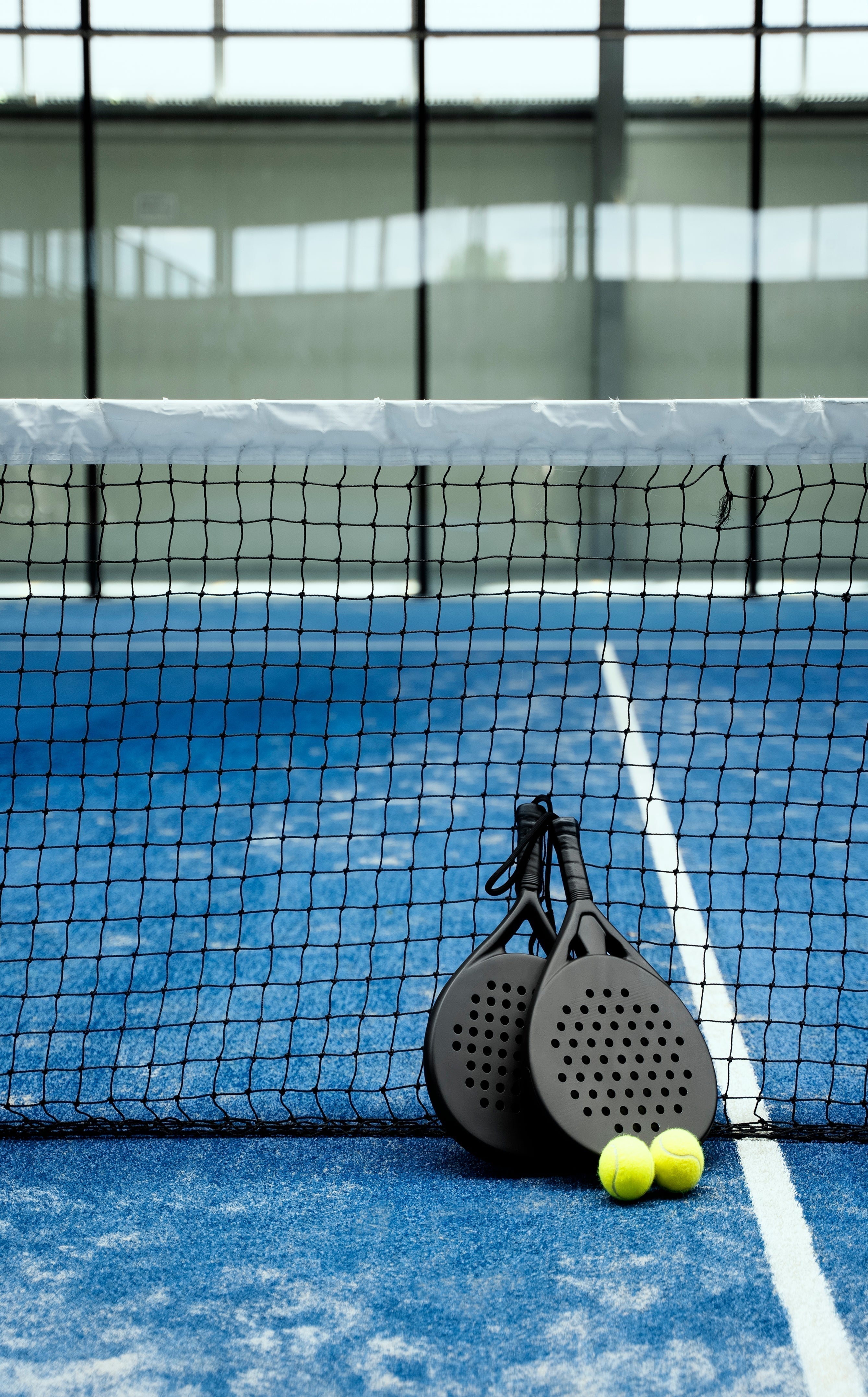 Two black padel rackets and two yellow balls resting against the net on a blue padel court.