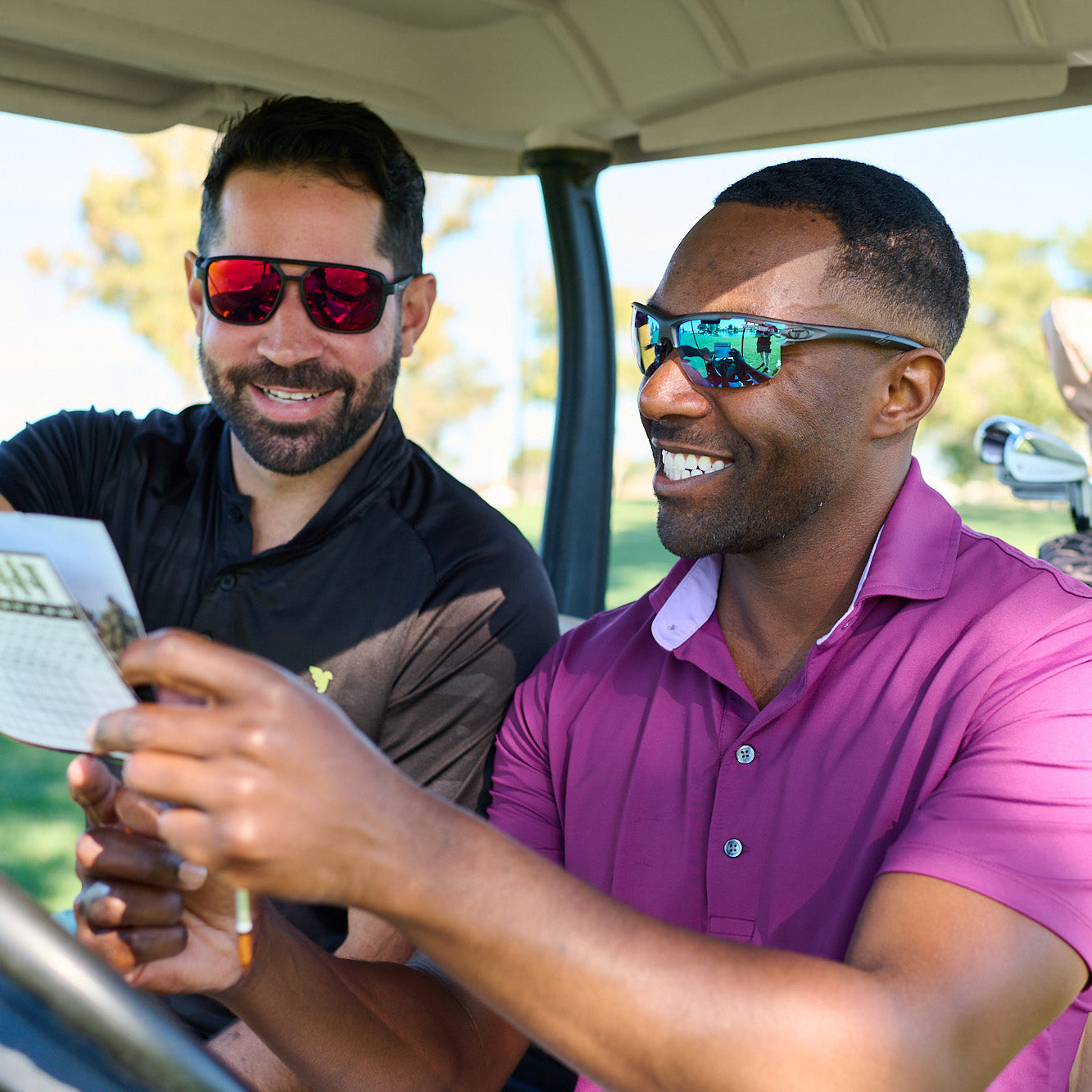 Two men wearing sunglasses sitting in a golf cart looking at a score card