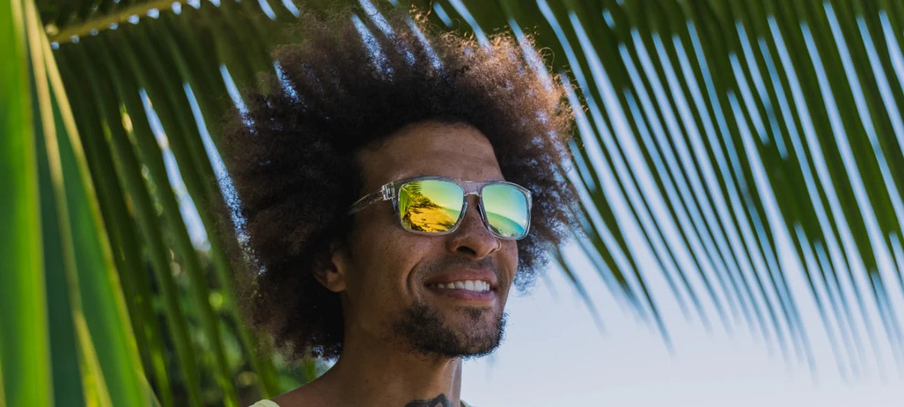 Man smiling and standing under a palm tree wearing Swick crystal clear sunglasses