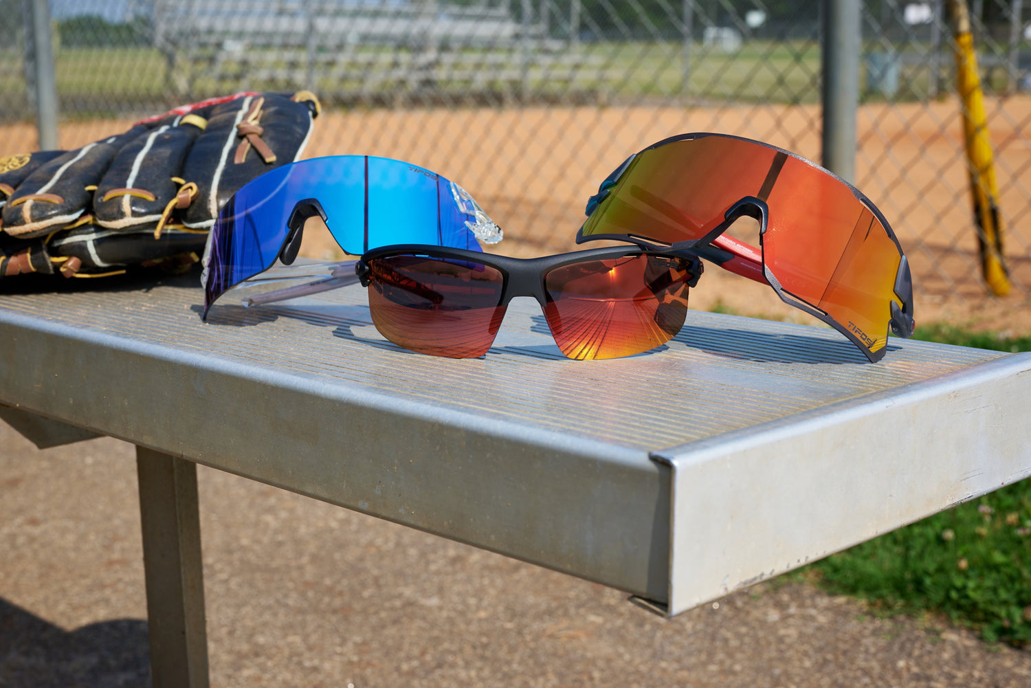 Three pairs of Tifosi baseball sunglasses on a bench beside a glove at a baseball field